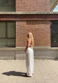 Vrouw met lang blond haar in witte broek en crop top staat zijwaarts op een zonovergoten stoep tegen een bakstenen muur.