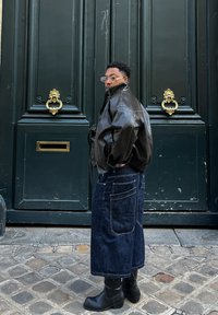 Jachetă bomber din piele maro, blugi din denim albastru cu picior larg și buzunare, și cizme negre până la glezne. Stă în fața unei uși verzi cu accesorii aurii.