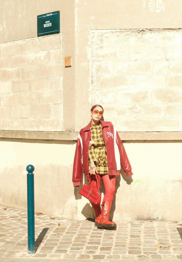 Person leaning against a beige wall, dressed in a red jacket, a yellow plaid dress, and red boots with tights, while holding a red handbag on a cobblestone street.