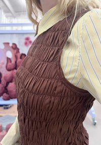Brun, struktureret, smocket tanktop lagdelt over en gul, kravebeklædt, stribet skjorte, hvilket skaber kontrast mellem farver og mønstre.