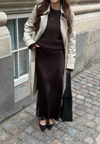 Μπεζ παλτό, καφέ πουλόβερ και σκούρα καφέ σατέν μακριά φούστα με άνοιγμα στο πλάι. Μαύρα σατέν γοβάκια με μυτερή μύτη και μαύρη τσάντα tote. Φόντο με πέτρες τουλάχιστον.