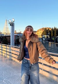 Jeune femme portant une veste à col en fourrure et un jean, faisant du patin à glace en plein air près d'une rambarde en bois dans un cadre urbain festif au coucher du soleil.