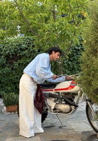Uma motocicleta branca com detalhes em vermelho está estacionada em uma calçada de pedra; uma pessoa a inspeciona, vestindo uma camisa azul clara e calças brancas, com um lenço na cabeça.