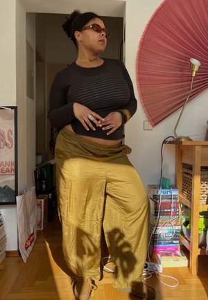 Mujer con gafas de sol, camiseta negra de manga larga, pantalones amarillos, de pie en el interior junto a una pila de libros y una decoración de pared en forma de abanico.