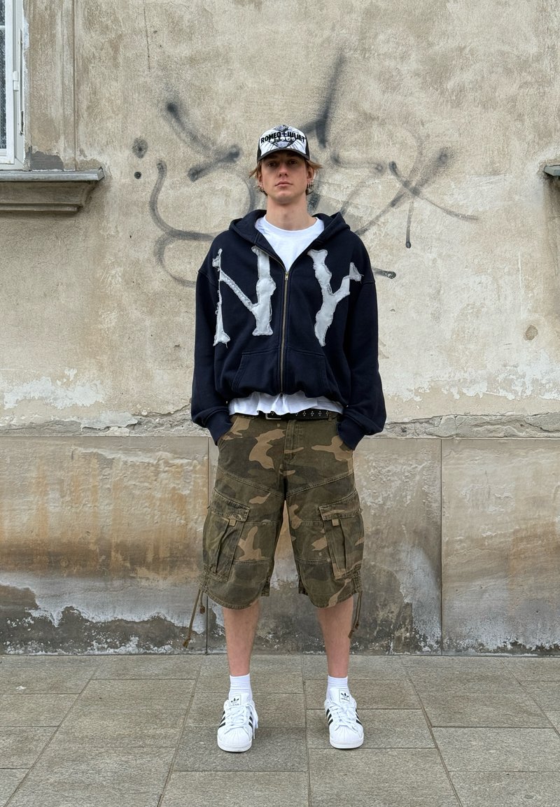 Jeune homme portant un sweat à capuche noir avec des lettres blanches, un short cargo camouflage, des baskets blanches et une casquette de baseball, debout devant un mur de graffiti.