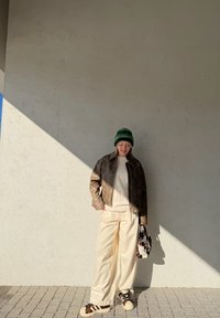 Casaco castanho com padrão, suéter creme, calças oversized creme, gorro verde e mala com padrão. Ténis com detalhes em preto e pele de Leopardo.