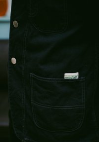 Svart lerret forkle med sølvfargede knapper, med en frontlomme og en etikett "Service World" brodert i grønt.