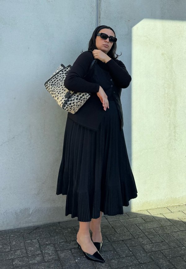 Mujer de pie contra una pared, vestida con vestido negro, cárdigan negro, gafas de sol negras, tacones negros y llevando un bolso tote negro con manchas blancas.