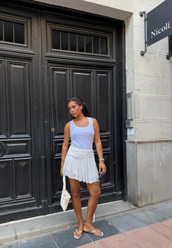 Femme en jupe blanche et débardeur bleu clair tenant un petit sac blanc et des lunettes de soleil, debout devant de grandes portes doubles noires.