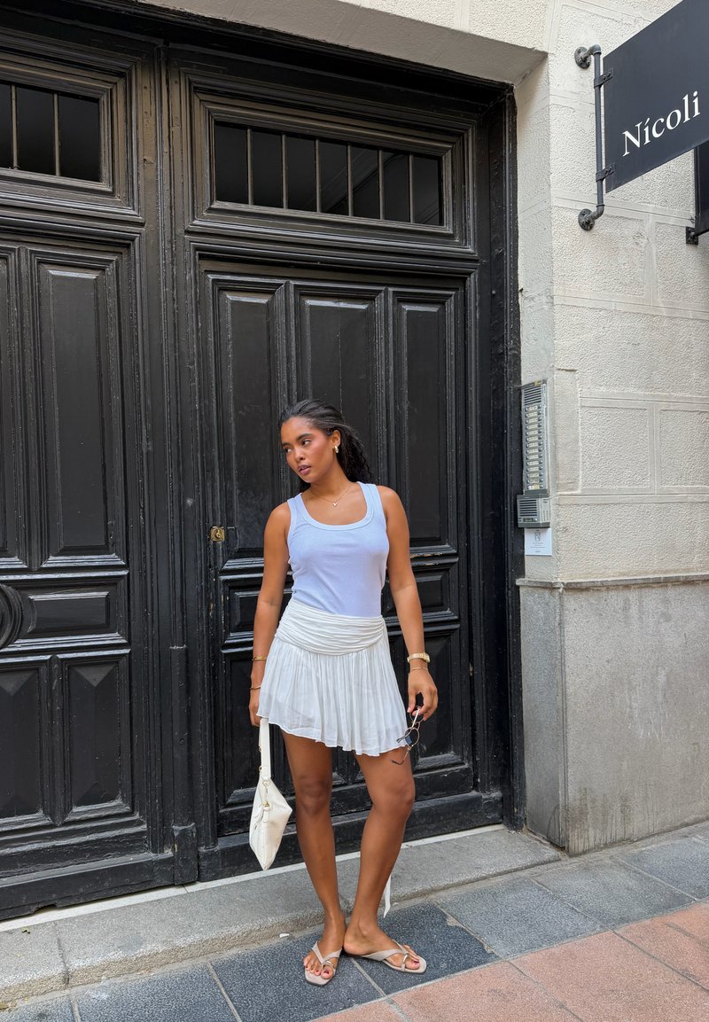 Femme en jupe blanche et débardeur bleu clair tenant un petit sac blanc et des lunettes de soleil, debout devant de grandes portes doubles noires.