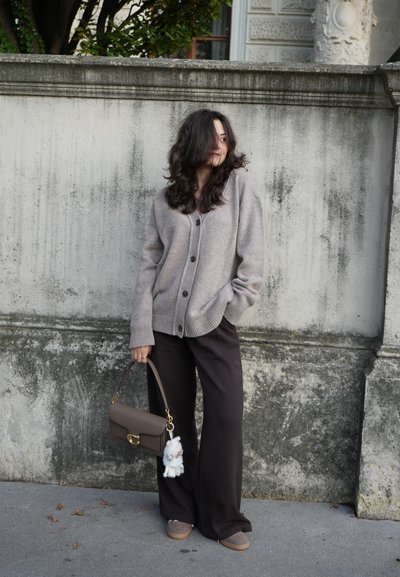 Cardigan beige con botones, combinado con pantalones de pierna ancha oscuros. Lleva un bolso marrón con un accesorio de peluche blanco. Texturas suaves y casuales.