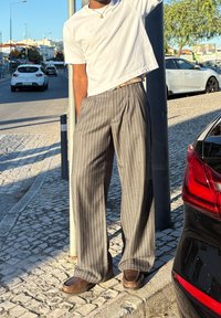 Pantaloni gri cu dungi subțiri, cu o croială lejeră, asortați cu un tricou alb scurt și încălțări maro. Stând pe o stradă pavata cu piatră cubică.