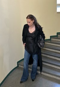 Manteau noir sur un haut noir orné de dentelle, associé à un jean bleu évasé et des escarpins noirs à bout pointu, tenant un sac à main noir.