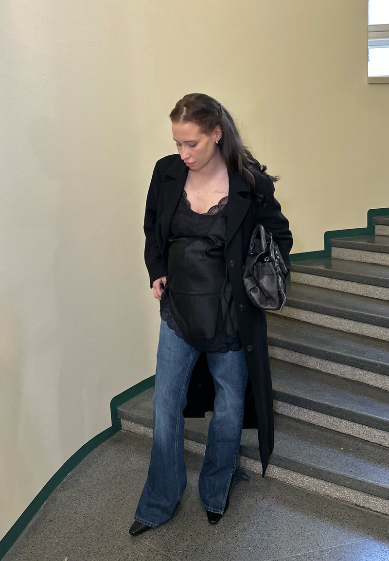 Črni plašč čez črn top z čipkasto obrobo, kombiniran z razširjenimi modrimi jeans hlačami in črnimi čevlji z ostrim prstom, drži črno torbico.