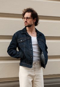 Giacca di jeans blu scuro con un taglio classico, chiusura a bottoni e tasche sul petto. Indossata sopra un top bianco, abbinata a pantaloni chiari.