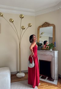 Robe rouge sans manches avec une jupe fluide, sac à main argenté et lampe sur pied moderne à arc doré. Cheminée en marbre avec des accents miroir.