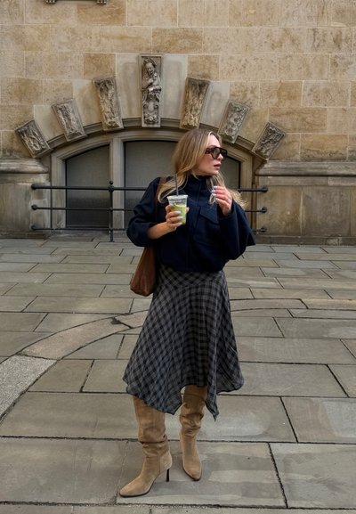 Mujer con gafas de sol y botas, sosteniendo una bebida verde, de pie en una calle empedrada con una pared decorativa de piedra detrás de ella.