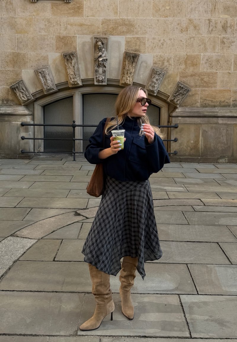 Frau trägt Sonnenbrille und Stiefel, hält ein grünes Getränk und steht auf einer gepflasterten Straße vor einer dekorativen Steinmauer.