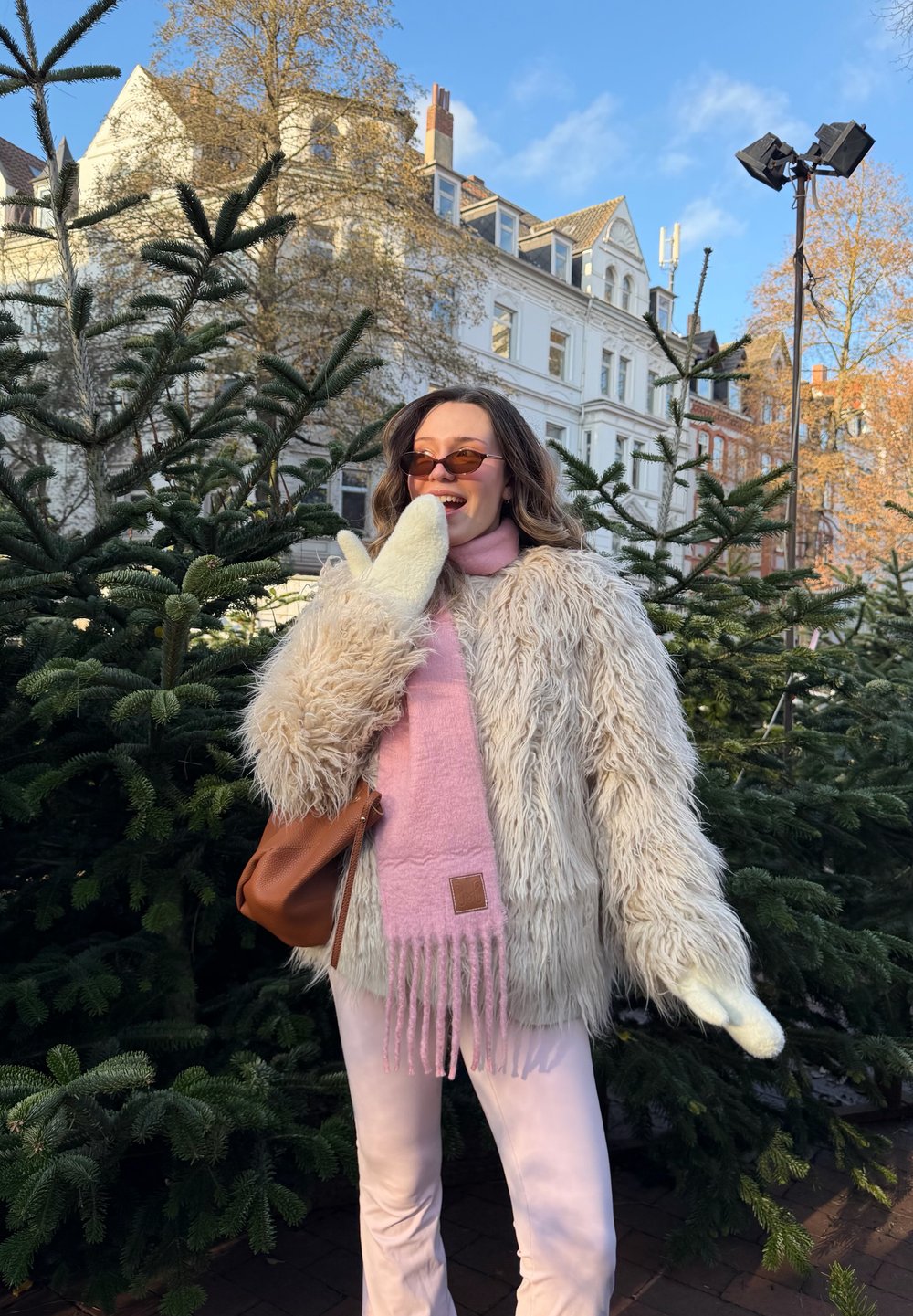 Furry beige coat, pink scarf, and white mittens. Brown handbag. Surrounded by evergreen trees with a backdrop of light-coloured buildings.