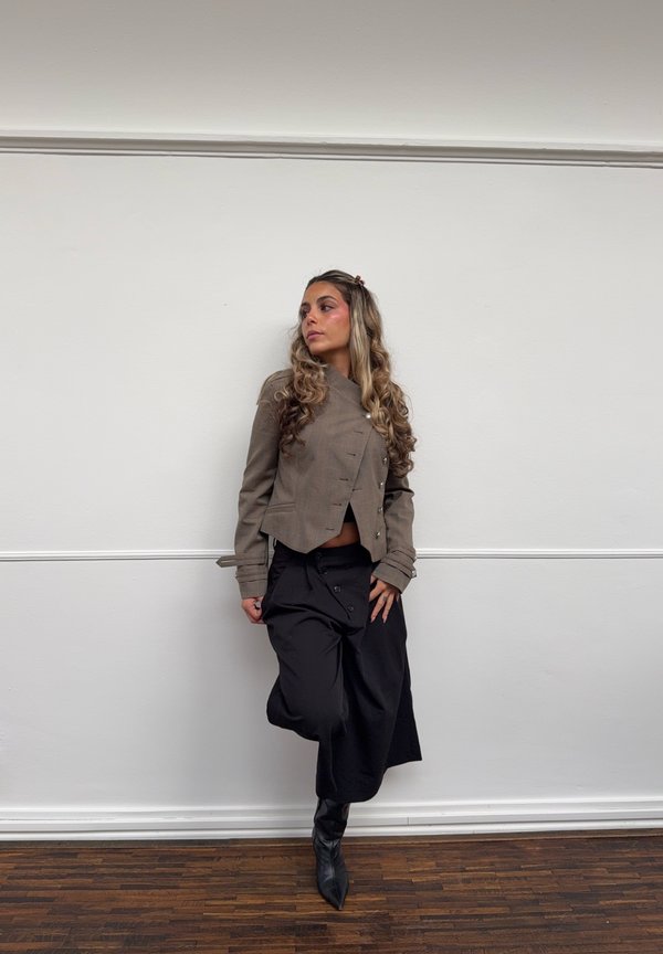 Femme aux longs cheveux bouclés appuyée contre un mur blanc, portant une veste courte marron, un pantalon noir à jambes larges et des bottes noires pointues sur un sol en bois.