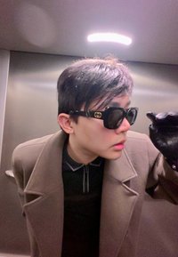 Svarta fyrkantiga solglasögon med guldfärgade logodetaljer, bärs med en brun kappa och en svart zip-piké, mot en metallisk hissbakgrund.