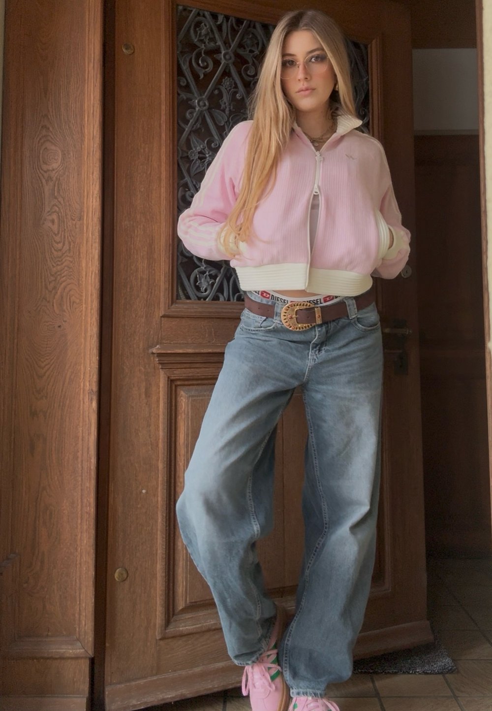 Jeune femme aux longs cheveux portant une veste rose, un jean bleu ample et des baskets roses, debout les mains dans les poches près d'une porte en bois.