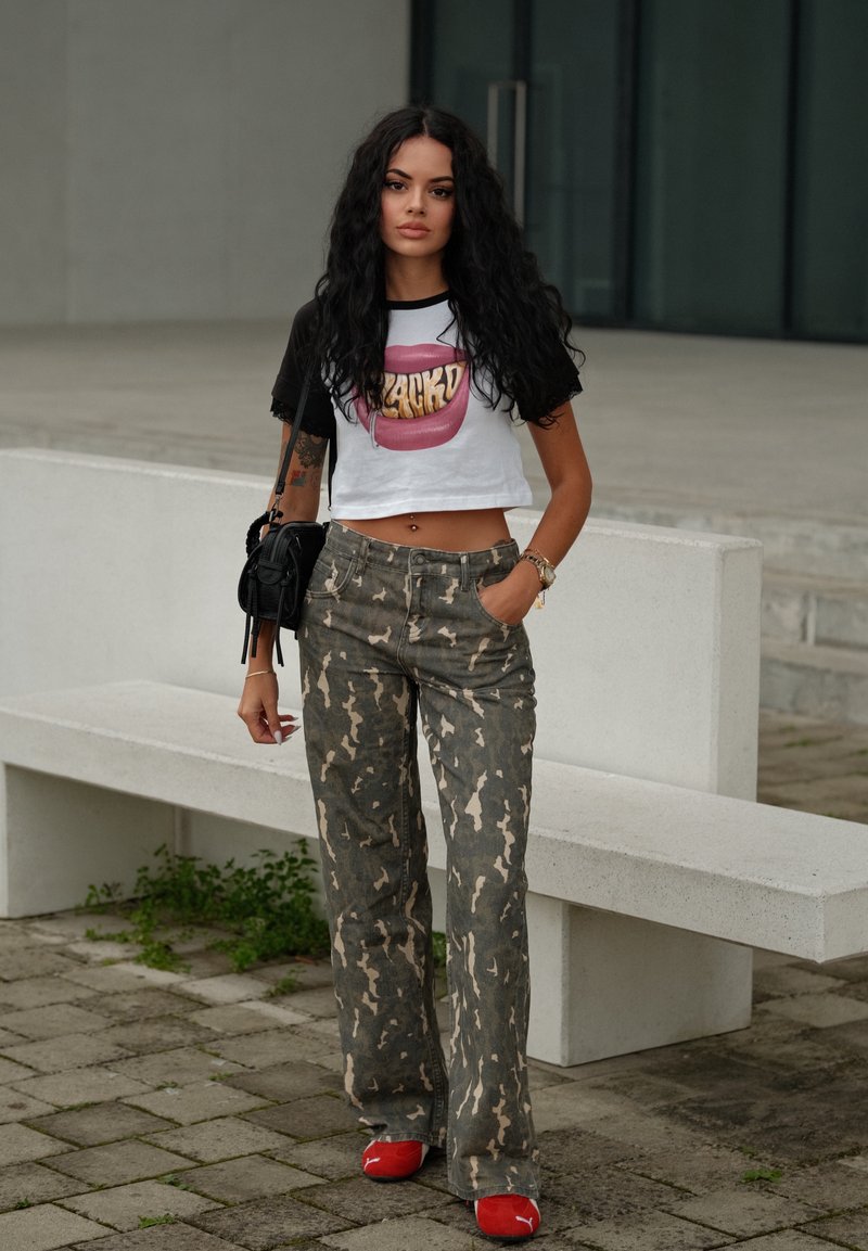 Crop top bianco con maniche nere con grafica di labbra rosa; pantaloni a gamba larga grigio mimetico; scarpe rosse; borsa nera; ambiente all'aperto.