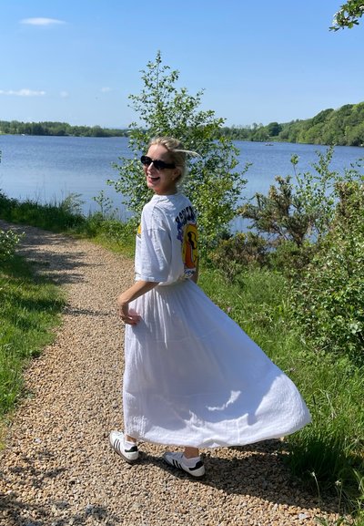 Vestido blanco y fluido con una camiseta gráfica de manga corta encima, combinado con gafas de sol negras y zapatillas blancas, ubicado junto a un sendero junto al lago.