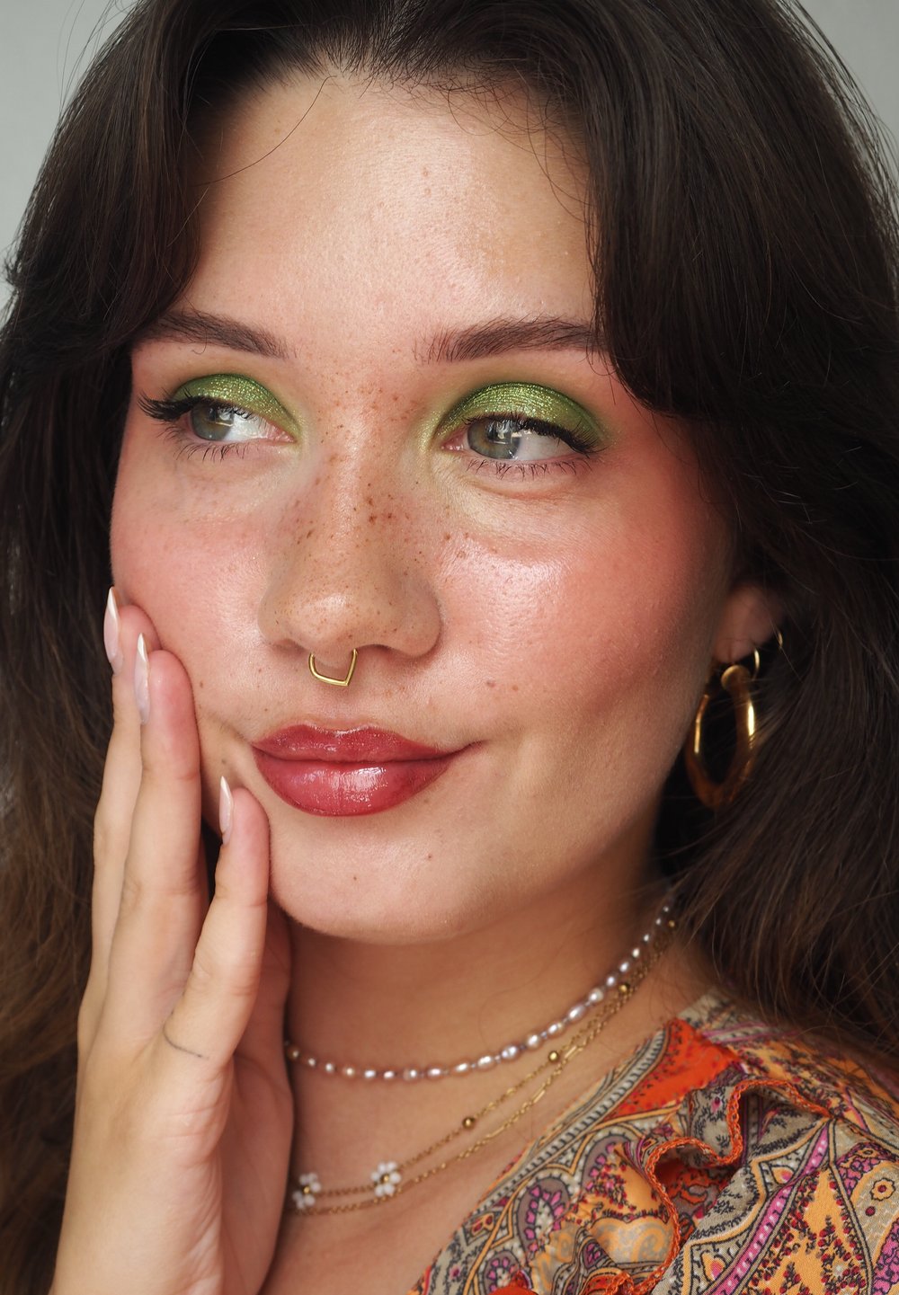 Nahaufnahme eines Gesichts mit grünem Lidschatten und Sommersprossen. Goldene Septum-Ringe, Kreolen und geschichtete Halsketten sind sichtbar. Die Haut wirkt dewy.