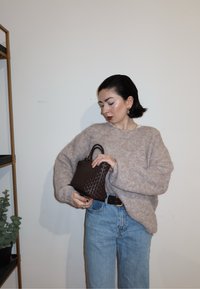 Femme aux cheveux courts et foncés ajustant un sac à main tissé foncé, portant un pull beige et un jean bleu clair, devant un mur blanc uni.