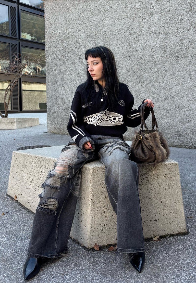 Black oversized sweater with graphic and stripe details, distressed gray jeans, black pointed heels, and a brown fur-accented handbag.