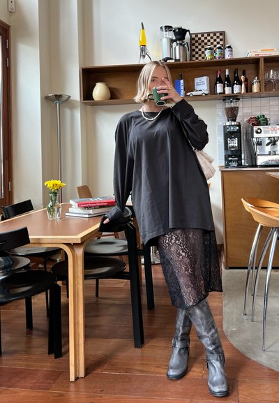 Camisa negra de manga larga, falda negra de encaje, botas grises hasta la rodilla. Accesorios incluyen un collar de capas y un bolso de hombro. Ambiente en un café interior.
