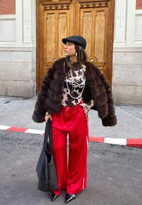 Giacca di pelliccia marrone, top con motivo nero e bianco, pantaloni sportivi rossi con strisce bianche, borsa nera a tracolla e scarpe nere a punta.