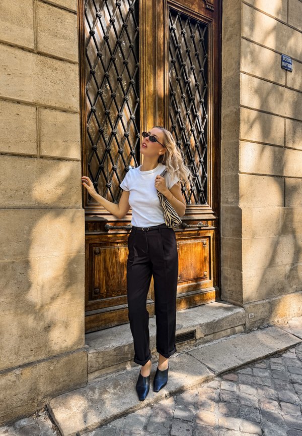 Woman in black pants and white shirt with sunglasses stands near large ornate wooden door on stone street.