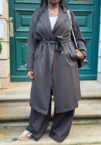 Mørkegrå, letvægts trench-coat med bælte, store knapper og sidelommer, parret med løse sorte bukser og beige sneakers.
