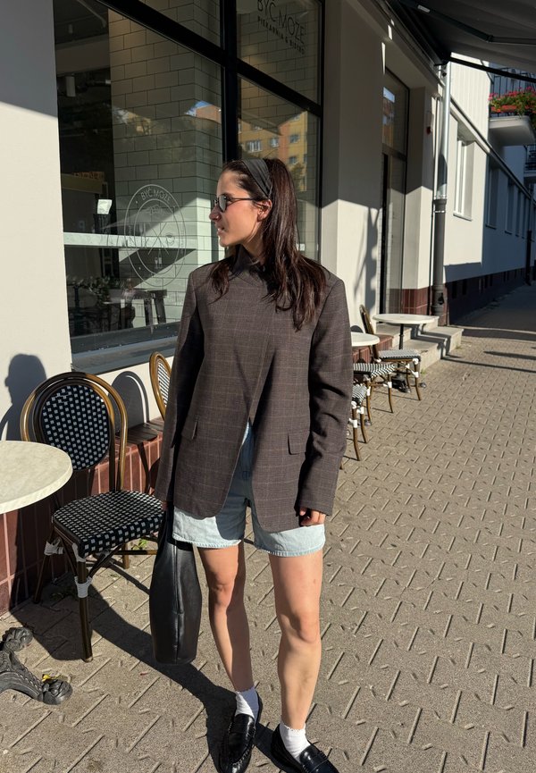 Femme portant un blazer foncé, un short en jean, des mocassins noirs et des lunettes de soleil tient un sac noir près des tables et des chaises d’un café en plein air.