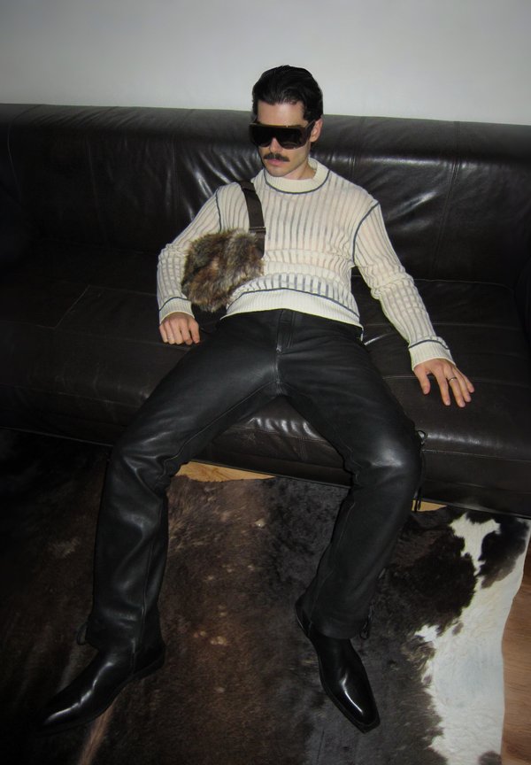 Pulover tricotat cu dungi crem, pantaloni negri din piele, cizme negre netede și geantă cu accente de blană, așezată pe o canapea întunecată din piele.