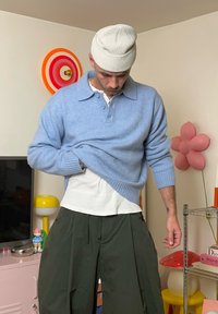 Lichtblauwe gebreide trui met kraag en drie knopen, over een wit overhemd gedragen, gecombineerd met loszittende donkergroene broek.