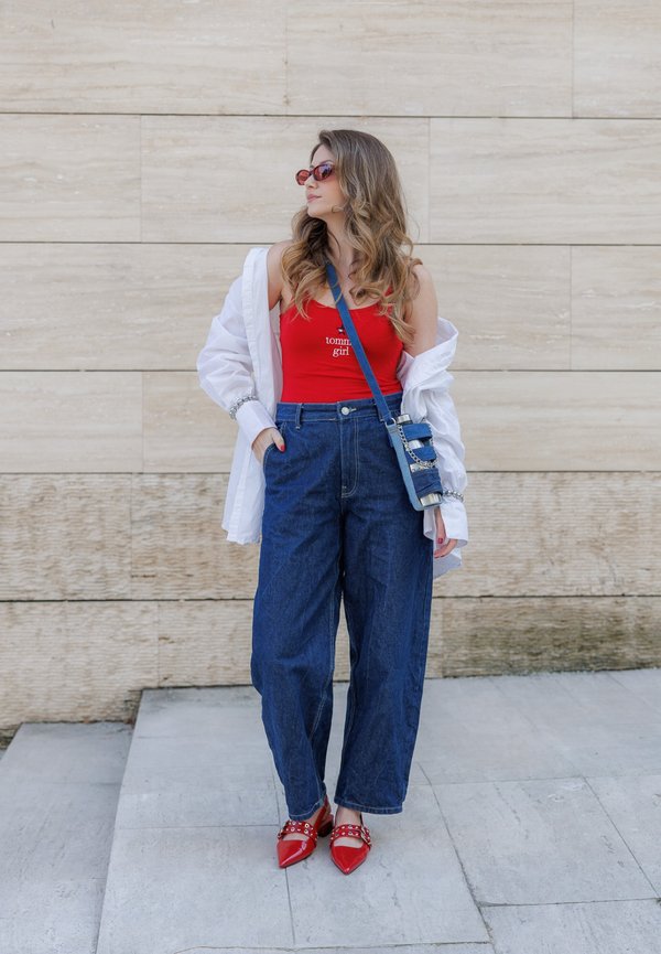 Une femme se tient contre un mur de pierre beige, portant un débardeur rouge, une chemise blanche ouverte, un jean à jambes larges, des chaussures rouges pointues, des lunettes de soleil et un sac bleu porté en bandoulière.