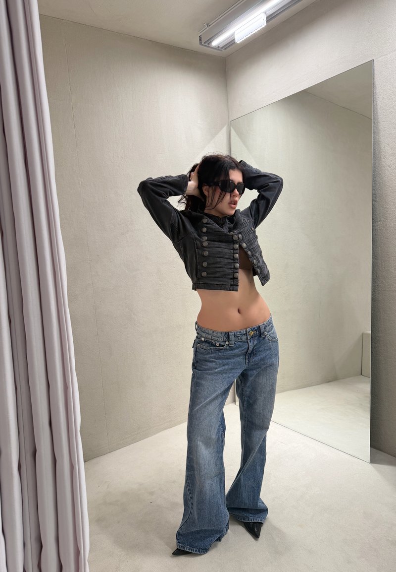 Woman in sunglasses wearing a cropped black jacket and wide-leg jeans poses with hands behind head in a minimal fitting room with large mirror.