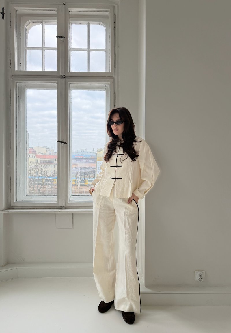 Femme portant une tenue blanche ample avec des garnitures noires et des lunettes de soleil, debout près d'une grande fenêtre dans une pièce minimaliste.