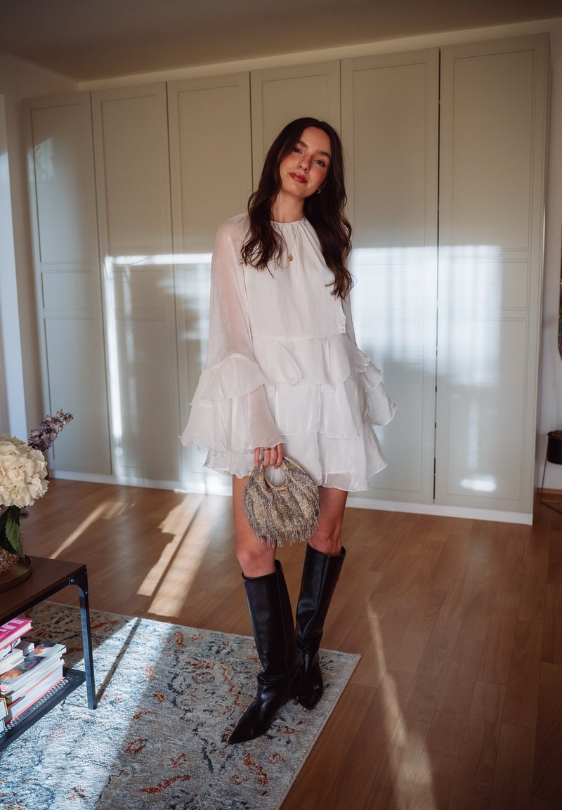 Femme en robe blanche à couches et bottes noires montantes tenant un sac à main texturé, debout dans une pièce ensoleillée avec un sol en bois.