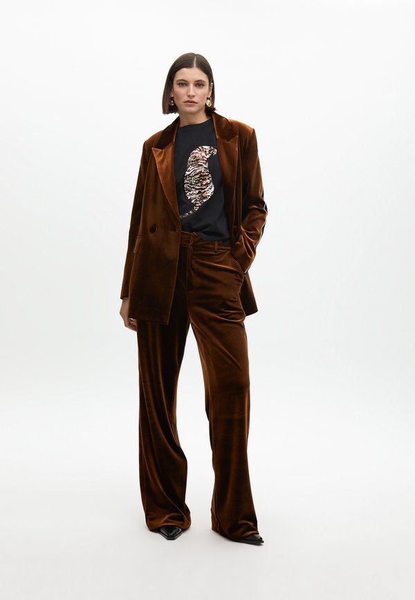 Braunes Samtanzug mit einem doppelt geknöpften Blazer und weiten Hosen. Schwarzes T-Shirt mit einem Pailletten-Tierdesign und spitzen schwarzen Schuhen.
