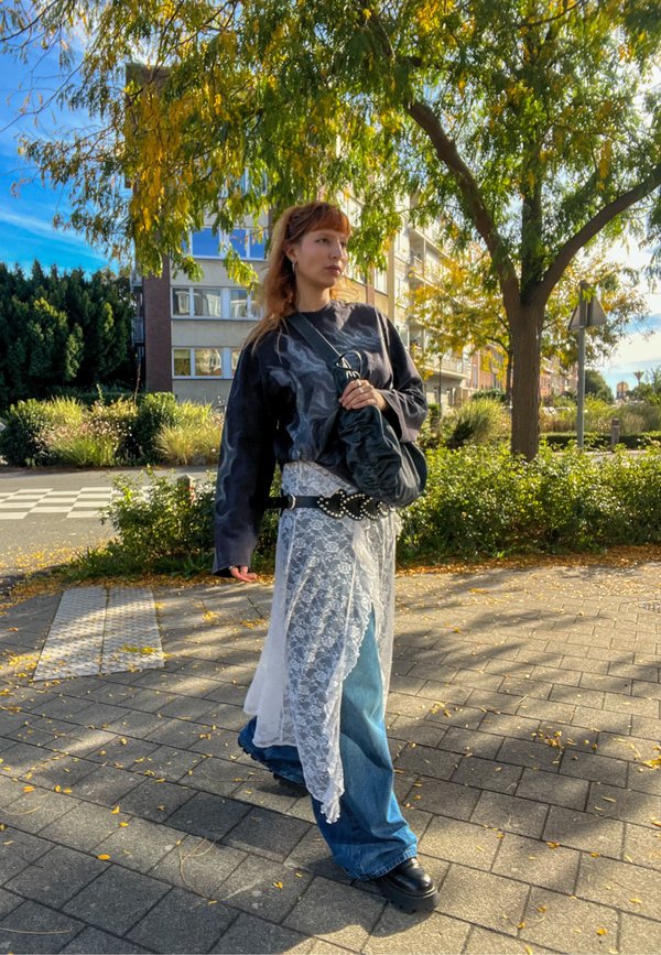Personne portant un pull sombre, une jupe en dentelle blanche par-dessus un jean bleu, des chaussures noires et portant un sac noir, marchant sur un trottoir ensoleillé avec des arbres et des bâtiments à proximité.