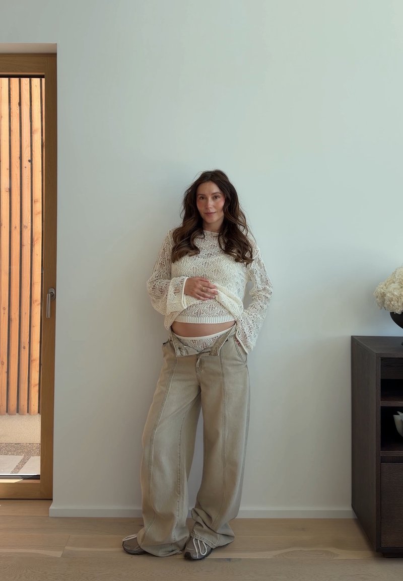 Femme enceinte portant un pull en dentelle blanc et un pantalon large beige se tient contre un mur blanc près d'une porte en bois et d'une armoire sombre.
