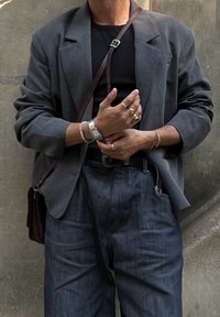 Siv križan blazer čez črno srajco, oblečen z temno denim hlačami. Dodatki vključujejo prstane in uro, vidna je tudi rjava torba čez ramo.