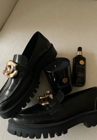 Paar zwarte chunky leren loafers met gouden kettingdetail naast een zwarte Precious Amber-kaars en huisparfum op beige stof.