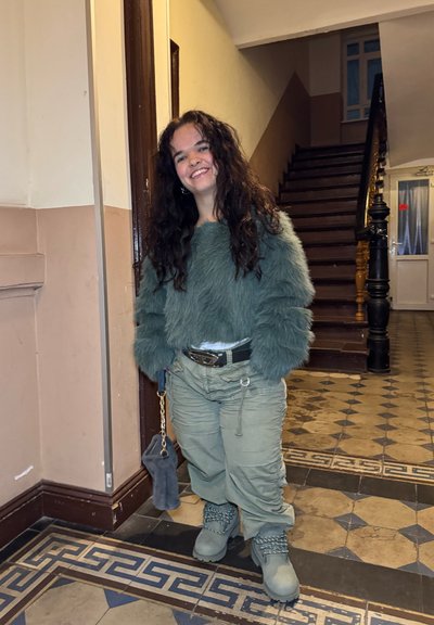 Suéter de pelo sintético verde, pantalones cargo olivo holgados, bolso gris con cadena y botas gruesas con detalles metálicos. Entorno interior con escaleras.