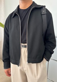 Crna jakna na zip s ovratnikom, uparen s crnom majicom i bež hlačama. Ima glatku teksturu i detalj na remenu za rame.