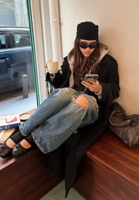 En person i en svart frakk og lue sitter på en benk, iført solbriller, holder en hvit kopp, med revne blå jeans og svarte sko.